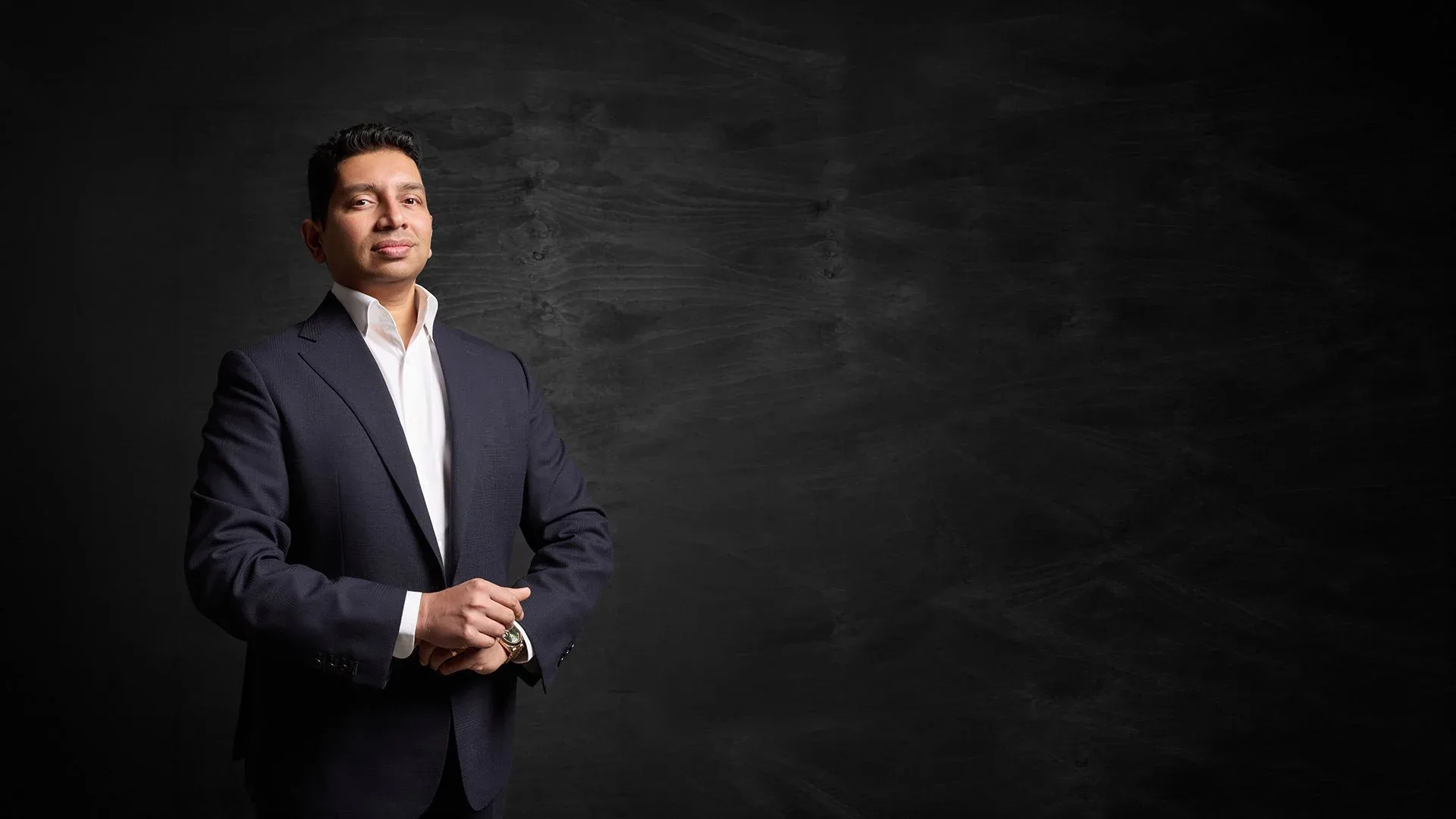Dr Tiv in a suit, hands clasped, lit against a dark textured studio backdrop